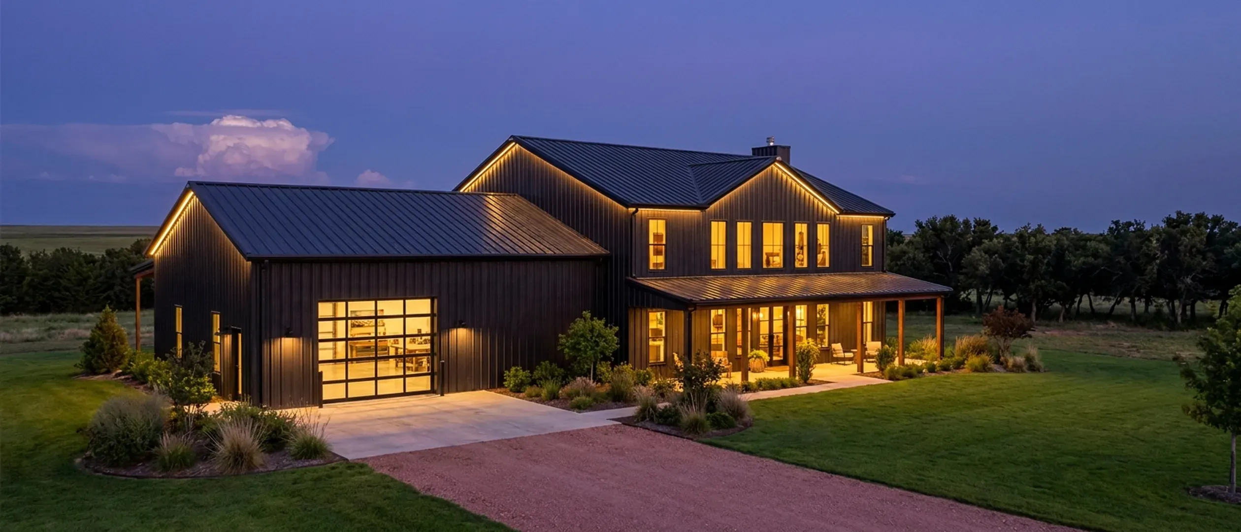 A barndominium at dusk with warm light glowing from every window and exterior lighting along the porch and roofline against a deep blue twilight sky.