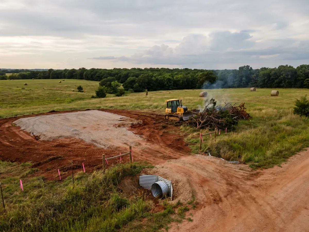 A dozer clearing cedar and brush on a rural parcel beside a compacted crushed-stone building pad, with red clay exposed and a culvert set at the driveway entrance.